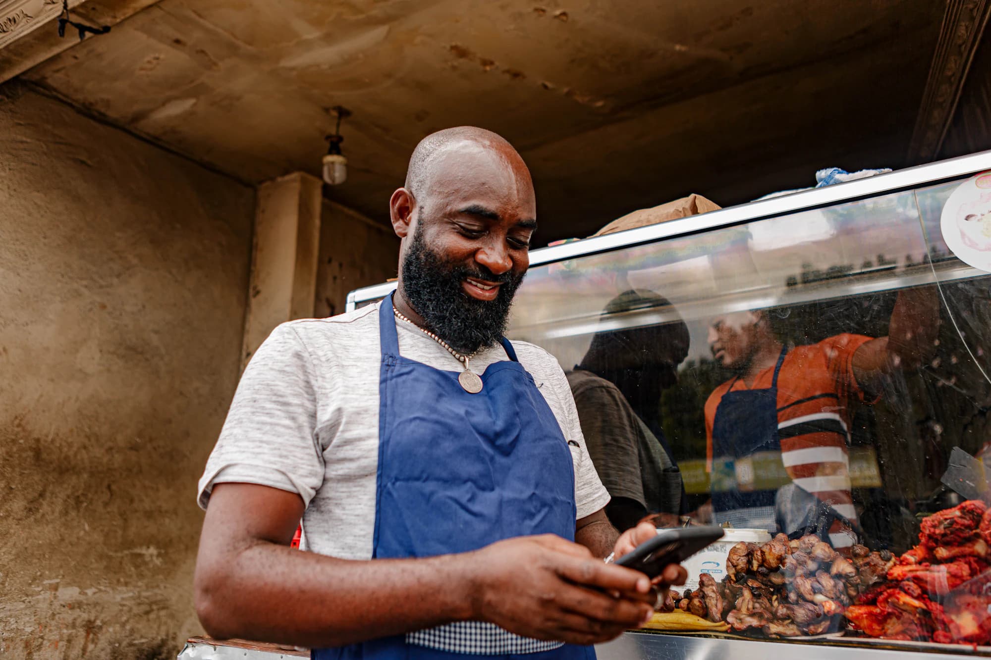 A food vendor in an apron smiling at his phone beside his display counter